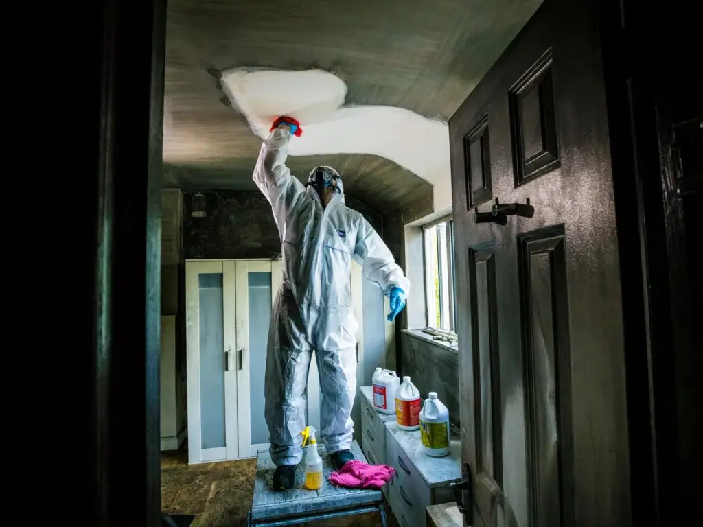 A technician in white coveralls cleaning soot and ash from a domestic property ceiling leaving a clear contras between the grey soot and white ceiling.