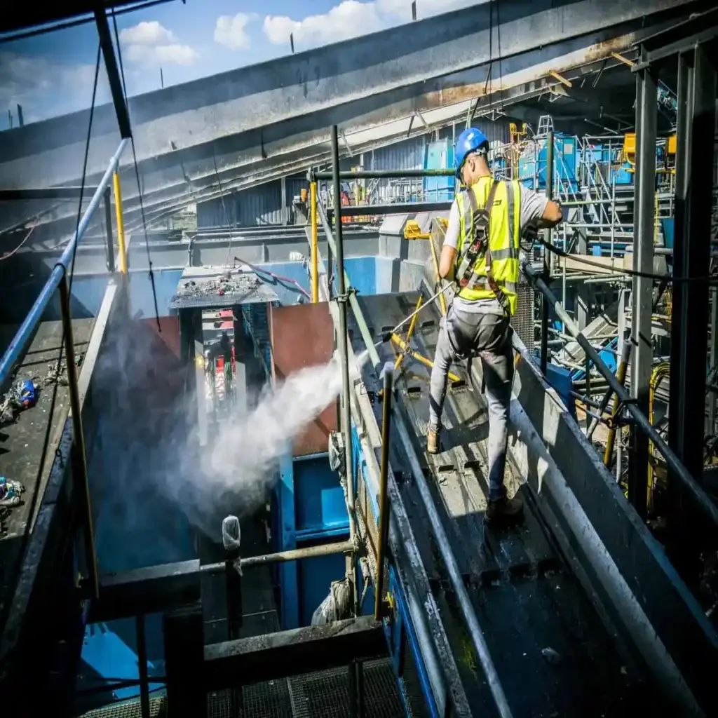 Technician cleaning an industrial facility with a jet washer on a walkway.