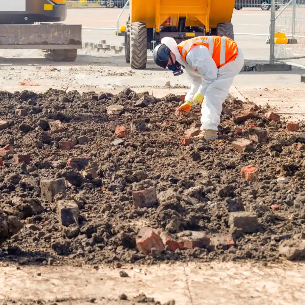 A technician in PPE removing asbestos from contaminated ground filled with bricks at a UK construction site